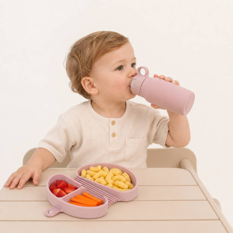 A toddler wearing a cream-colored henley shirt sits at a light wood table, drinking from a dusty pink reusable bottle. In front of the child is a matching pink divided meal tray containing yellow pasta, red strawberries, and orange carrot sticks.